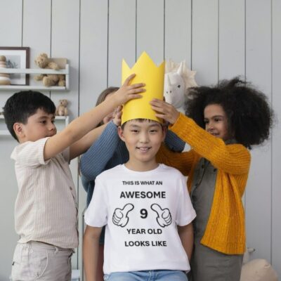 Boy wearing a T-shirt that says "This is what a 9 year old looks like" with 3 other kids putting a yellow crown on his head