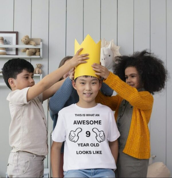 Boy wearing a T-shirt that says "This is what a 9 year old looks like" with 3 other kids putting a yellow crown on his head